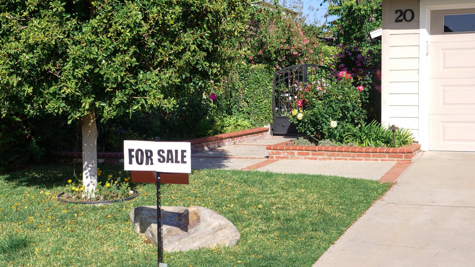 Front view of a house with a garden and 'For Sale' sign in bright daylight.